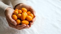 Hands holding fresh orange kumquats on white background, ideal for food and wellness marketing materials.