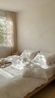 Serene bedroom interior with unmade bed featuring white linens, natural sunlight streaming through sheer curtains, minimalist decor, and wooden flooring for home design inspiration.