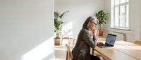 Senior businesswoman with gray hair working on laptop at wooden desk in bright modern office with plants and natural light streaming through windows.
