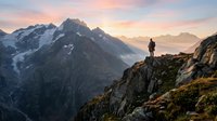 Silhouette of hiker standing on rocky mountain ridge at sunrise with snow-capped peaks and misty valleys in background. Adventure and outdoor exploration.