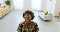 Middle-aged woman with dark skin tone wearing traditional patterned clothing and head wrap, smiling at camera in bright, minimalist living room with natural light, wooden furniture and neutral decor.
