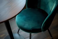 Elegant emerald green velvet dining chair with curved backrest and black metal legs against wooden table, modern interior design element in moody lighting.