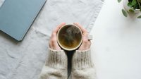 Hands in cozy knit sweater holding a steaming cup of herbal tea on a white table with linen cloth and tablet nearby.
