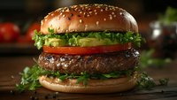 Fresh juicy beef burger with lettuce, tomato, and avocado slices on sesame bun against dark wooden background. Close-up view shows texture and layers.