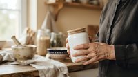 Senior woman with wrinkled hands holding white ceramic mug in warm kitchen with natural light, ideal for lifestyle and home decor content.