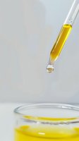Glass dropper with yellow liquid suspended above laboratory glassware against white background, capturing moment before drop falls into solution.