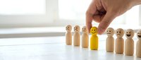 Wooden peg dolls with drawn faces in a row, one yellow figure highlighted by hand, representing customer satisfaction, employee recognition, or standing out from crowd.