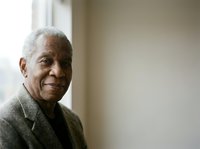 Senior African American man with gray hair wearing olive green blazer and black shirt, smiling warmly while looking at camera near window with natural light.