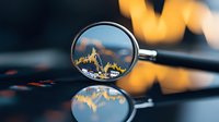 Magnifying glass showing financial chart data with golden graph line against blurred trading screen background for market analysis.