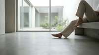 Barefoot person in linen pants relaxing on concrete steps in a minimalist modern home with large windows and courtyard view.