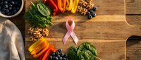 Pink ribbon surrounded by healthy foods including blueberries, spinach, bell peppers, carrots and walnuts on wooden cutting board for cancer awareness.