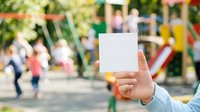 Child's hand holding blank white card at playground with blurred kids playing in background, suitable for advertising, marketing messages, or promotional campaigns.