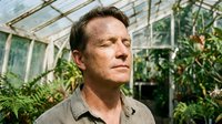 Middle-aged Caucasian male botanist or horticulturist in greenhouse surrounded by green plants and foliage, wearing casual work shirt, suitable for agriculture and gardening industry content.