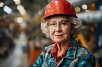 Senior female engineer wearing red hard hat, plaid shirt and glasses smiling confidently in industrial setting. Professional portrait showcasing diversity in workforce.