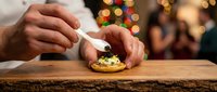 Caucasian male chef carefully placing caviar on a blini appetizer on a rustic wooden board with festive bokeh lights in background.
