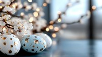 Easter eggs decorated in light blue and white with gold pattern near blooming cherry branches against blurred bokeh lights creating festive spring atmosphere.