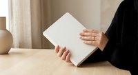 Woman in black sweater reading blank white book at wooden table in cozy home interior with neutral decor and soft natural lighting.