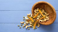 Wooden bowl with mixed nuts and dried fruits spilling onto blue wooden background for healthy snacking and cooking ingredients.