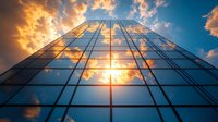 Modern glass office building facade reflecting dramatic sunset sky with golden clouds, viewed from below looking up. Architectural perspective symbolizes success and growth.