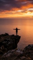 Silhouetted figure standing on rocky cliff with arms outstretched overlooking calm ocean during golden sunset, conveying freedom, achievement, and contemplation for wellness and lifestyle concepts.