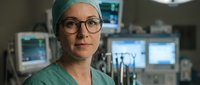 Female surgeon with glasses in green scrubs and surgical cap standing in modern operating room with medical equipment and monitors in background.