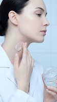 Young woman applying moisturizer cream to neck from glass jar. Clean skincare routine, anti-aging treatment for healthy glowing skin in white bathrobe.