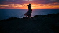 Silhouetted woman in flowing dress standing on rocky coastline at sunset, watching ocean horizon with dramatic golden and purple sky reflecting on water.
