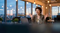 Young Hispanic businesswoman celebrating success with raised hands in modern office during evening hours with city skyline view and warm lighting.