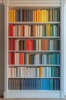 Books arranged by color spectrum on white wooden bookshelf creating rainbow pattern from yellow through red to blue. Classical library interior design element.