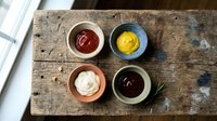 Four ceramic bowls with ketchup, mustard, mayonnaise and BBQ sauce arranged on a rustic wooden table, top view, food styling.
