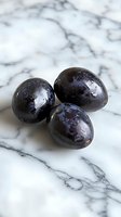 Fresh ripe dark plums with water drops on glossy skin, arranged on white marble surface. Natural lighting enhances fruit texture and marble veining.