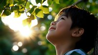 Young Asian boy looking up at sunlight through green leaves with joyful expression. Natural bokeh effect creates dreamy atmosphere in outdoor setting.