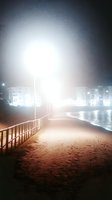 Misty night scene along sandy beach boardwalk with wooden railing, illuminated by bright street lamps casting ethereal glow, waterfront buildings visible in background.