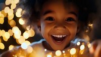 Young African American girl with curly hair smiling joyfully while holding warm glowing string lights, creating magical bokeh effect for holiday celebrations.