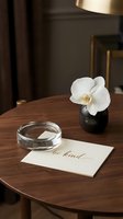 Elegant wooden table with white orchid in black vase, glass bowl, and be kind calligraphy card in warm interior setting.