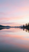 Serene alpine lake at sunrise with pink and purple sky reflecting on calm water, evergreen trees lining shoreline, mountains in background.
