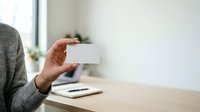 Young adult with medium skin tone holding blank white business card at wooden desk with notebook and smartphone, suitable for corporate branding and professional networking concepts.