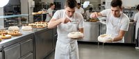 Male bakers in white uniforms and aprons prepare fresh bread and pastries in a professional kitchen bakery setting with equipment and colleagues working.