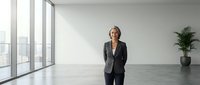 Mature woman in business attire standing in modern empty office space with large windows and potted plant, suitable for corporate and professional concepts.