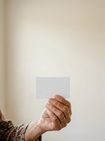 Senior adult hand holding blank white card against neutral beige wall background, suitable for mockups, business messaging, and promotional campaigns.