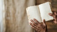 Senior adult with wrinkled hands holding open aged book with blank pages against neutral background. Suitable for education, literature, reading, nostalgia, and heritage concepts.