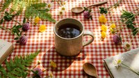 Rustic ceramic mug with hot coffee on red checkered tablecloth surrounded by autumn leaves, ferns and wooden spoons for cozy seasonal morning beverage concept.