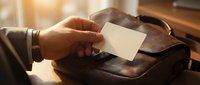 Professional businessman hand holding blank white business card over leather briefcase on wooden desk in warm office lighting for corporate branding mockup.