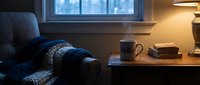 Cozy winter evening corner with steaming mug on wooden nightstand, knit blanket on armchair, warm lamp glow, and snowy window view.