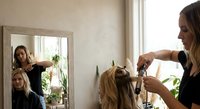 Professional hairstylist cutting blonde woman's hair in modern salon with mirror reflection and natural lighting for beauty services.