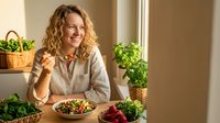 Happy blonde woman enjoying healthy salad meal at home kitchen table surrounded by fresh green plants and vegetables for wellness lifestyle.