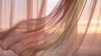 Flowing sheer fabric in soft pink and gold tones with warm sunlight creating elegant waves and movement through translucent material.