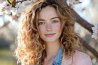 Young Caucasian woman with natural curly blonde hair and gentle smile posing near blooming spring cherry tree branches in soft evening sunlight.