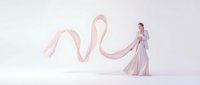 Young woman in flowing white dress dancing with long pink silk ribbon against soft pastel background. Graceful movement and ethereal atmosphere for wellness concepts.