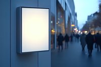 Blank illuminated lightbox on city street wall with blurred people walking at dusk, mockup for advertising and promotional content display.